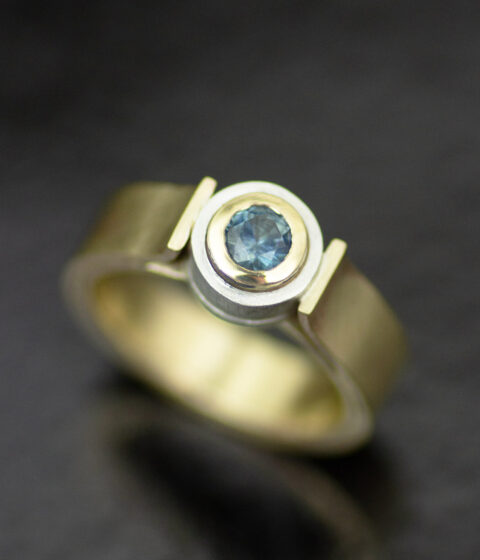 A teal montana sapphire set in a silver bezel setting, on a gold ring band.